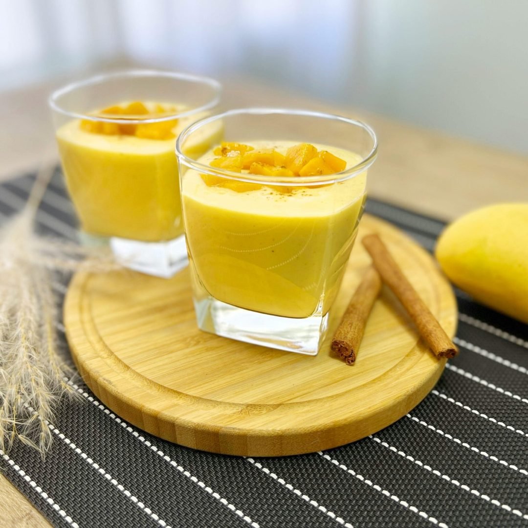 Two glasses of creamy mango lassi garnished with diced mango and cinnamon sticks.